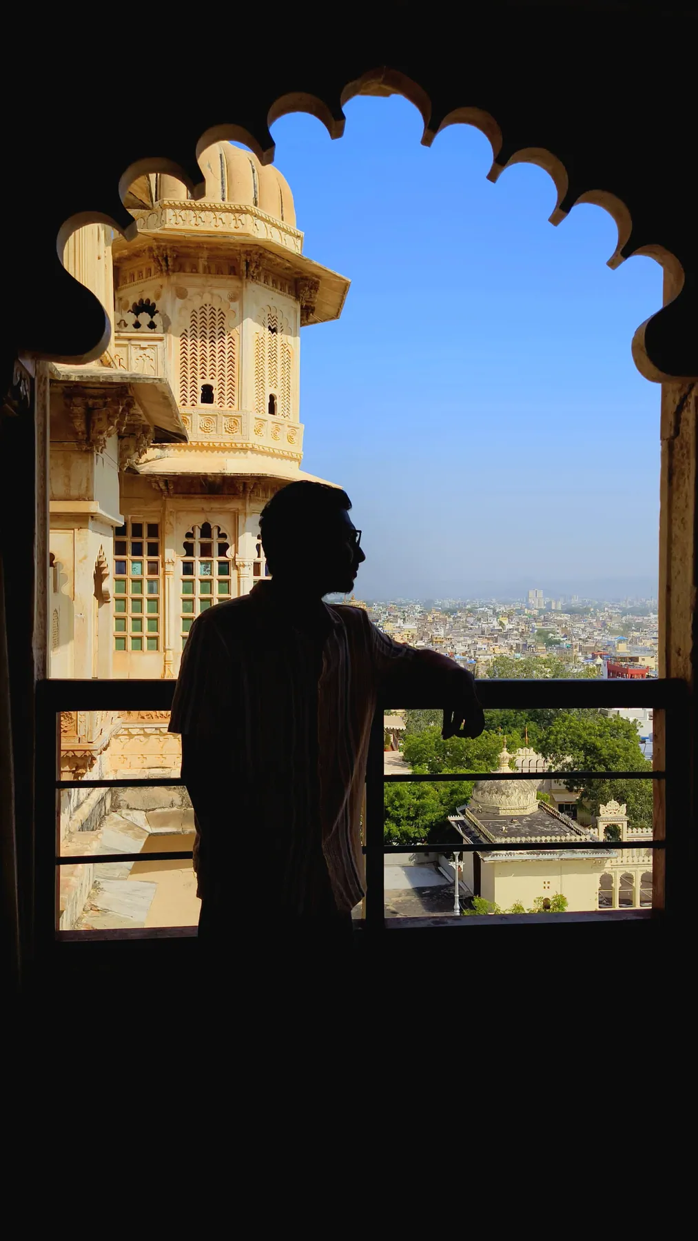 Gantavya in silhouette at a scalloped palace window, looking out over a city