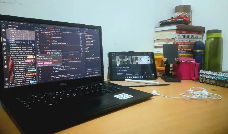 A desk at night: a laptop open on code, a tablet, earphones, a water bottle and a stack of books.