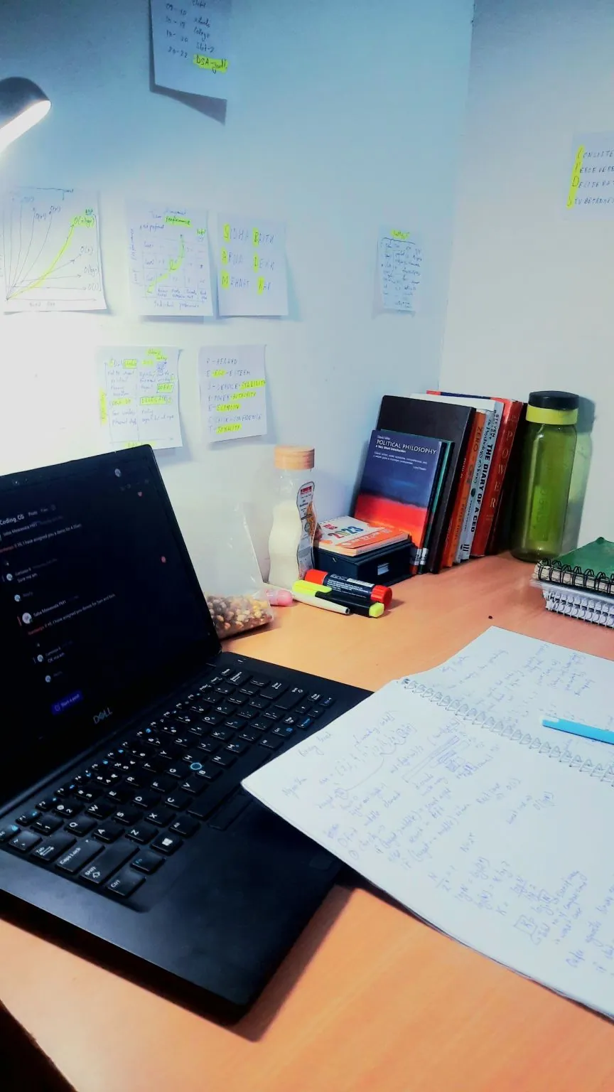 A desk under a lamp: a laptop, a notebook full of handwritten working, books, and sticky notes of plans on the wall.